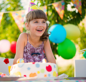 Little girl laughing at birthday party, wearing birthday hat and sitting in front of birthday cake with balloons